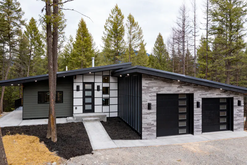 A Built Right modern home in the pines by day: a stacked-stone wall, a grid of glass at the entry and a long low roofline under tall ponderosa, Modern Living Montana Built